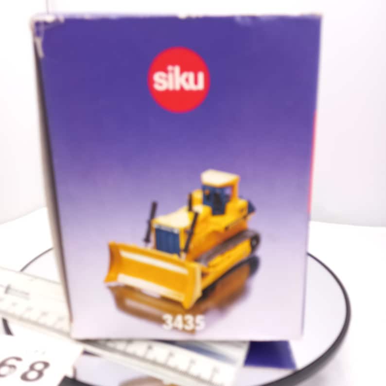 May include: A yellow and black toy bulldozer with a front blade, tracks, and a cab, presented in a box with the brand name "siku" and the number "3435". The box has a purple background.