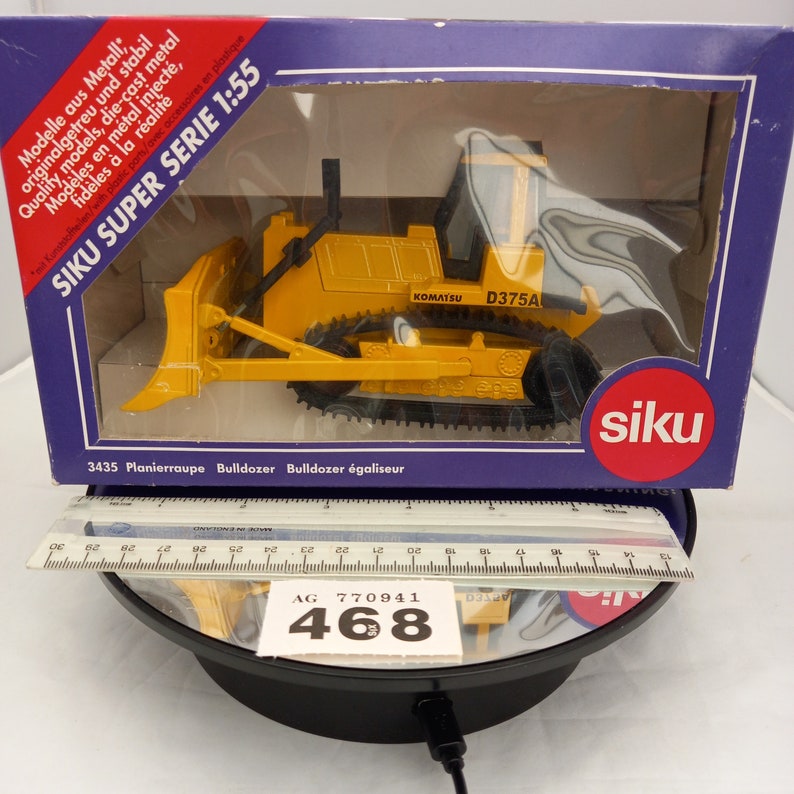 May include: A yellow Komatsu D375A bulldozer model in a box. The model is die-cast metal and plastic, with black tracks and a front blade. The box is blue and white, with text in multiple languages, including "SIKU SUPER SERIE 1:55".