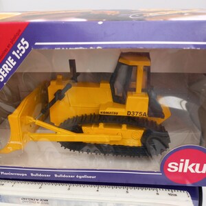 May include: A yellow Komatsu D375A bulldozer model, scale 1:55, in a clear plastic display case. The model features black tracks and a front blade. The brand name "siku" is visible on the packaging.