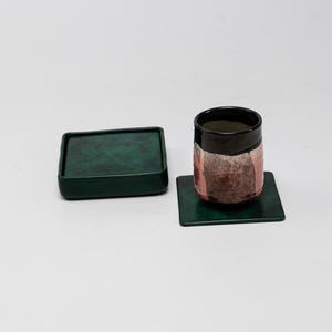 May include: A ceramic tumbler with a black rim and a pink and brown design, resting on a square green coaster. A matching green square tray sits nearby. The items are on a white background.