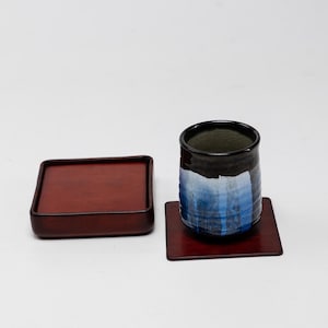 May include: A Japanese tea set featuring a black and blue ceramic cup with a matching coaster, and a square, red lacquered tray. The cup has a unique blue and white painted design. The set is displayed on a white background.