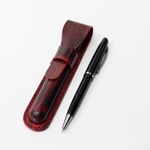 May include: A black pen with silver accents rests beside a burgundy leather pen case. The pen case has a flap closure and decorative stitching around the edges. The pen is sleek and modern.