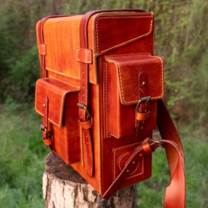 Puede incluir: Un bolso de cuero color coñac, estructurado y rectangular, con múltiples bolsillos exteriores con cierre de solapa, hebillas de metal color latón y costuras visibles. El bolso tiene una orientación vertical con correas de cuero ajustables.