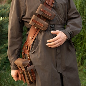May include: A brown leather utility belt with multiple pouches and holsters. The belt features a shoulder strap and is worn over a dark brown coat. The pouches are secured with leather straps and metal hardware.