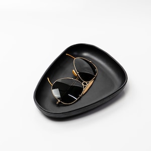 May include: Black sunglasses with gold-colored frames rest in a black, triangular-shaped tray. The sunglasses have dark lenses and a classic aviator design. The tray has a smooth, matte finish.