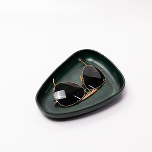 May include: A dark green, irregularly shaped tray holding a pair of sunglasses. The sunglasses have black lenses and gold-colored frames. The tray is empty except for the sunglasses.