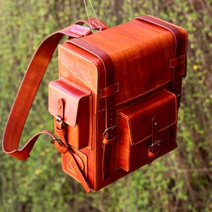 May include: A handcrafted, rectangular leather backpack in a rich, reddish-brown hue. The bag features two front pockets with buckle closures, a top handle, and adjustable shoulder straps. The stitching is visible, and the bag appears to be suspended against a blurred green background.