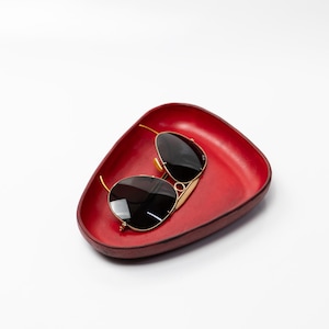 May include: A pair of sunglasses with black lenses and gold frames rests in a red, triangular tray. The tray has a smooth, glossy finish and a slightly raised edge. The sunglasses are positioned diagonally within the tray.