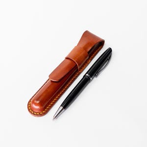 May include: A black pen with silver accents rests beside a brown leather pen case. The pen case is rectangular with a flap closure and a stitched border. The pen and case are on a white background.