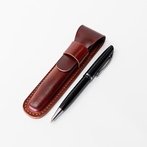 May include: A black pen with silver accents rests beside a brown leather pen case. The pen case is rectangular with a flap and a loop closure. The pen and case are set against a white background.