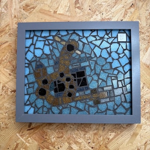May include: A mosaic artwork in a gray frame. The mosaic features a design with gold, black, and blue tiles. The background is a light blue, and the frame is gray. The artwork has a modern, abstract style.
