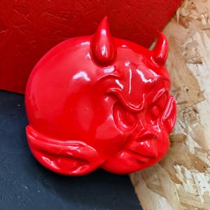May include: A glossy, bright red devil head sculpture with prominent horns, a furrowed brow, and a mischievous expression. The sculpture is a detailed, three-dimensional artwork, likely for decorative purposes.