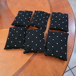 May include: Six black fabric bean bags with white polka dots are arranged on a wooden table. The bean bags are rectangular and appear to be filled with a soft material. The table has a warm, brown finish.