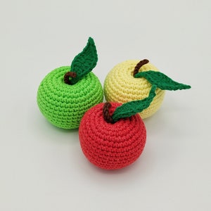 May include: Three crocheted apples in vibrant colors: green, yellow, and red. Each apple has a green leaf and a brown stem. The handmade apples are arranged on a white surface.