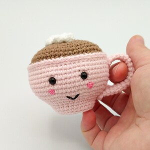 May include: A pink crocheted coffee cup with a brown crocheted coffee inside and a white crocheted cream on top. The cup has a happy face with black eyes and a pink smile.