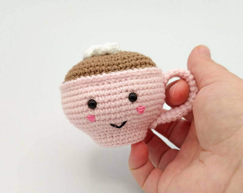 May include: A pink crocheted coffee cup with a brown crocheted coffee inside and a white crocheted cream on top. The cup has a happy face with black eyes and a pink smile.