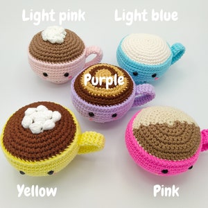 May include: Five crocheted coffee cup plushies in different colors. The cups are light pink, light blue, purple, yellow, and pink. Each cup has a brown crocheted rim and a white crocheted top. The cups have cute faces with black eyes and a small mouth.