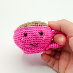 May include: A pink crocheted coffee cup with a brown crocheted top and a smiling face. The cup has black eyes and pink cheeks.