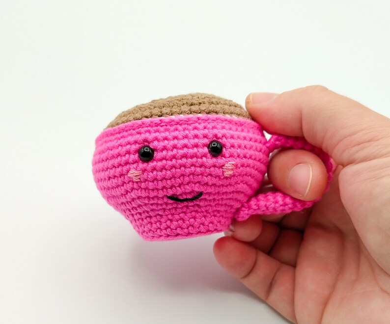 May include: A pink crocheted coffee cup with a brown crocheted top and a smiling face. The cup has black eyes and pink cheeks.