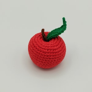 May include: A crocheted red apple with a brown stem and a green leaf. The handmade fruit is round and textured, perfect for a child's toy or decorative item. The apple is set against a plain white background.