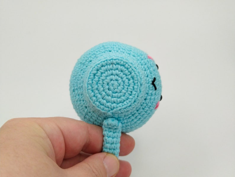 May include: A light blue crocheted stuffed toy shaped like a teacup with a pink and black face. The toy has a small handle.