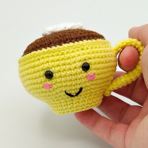 May include: A crocheted yellow coffee cup with a brown top and a white cream swirl. The cup has a happy face with black eyes and pink cheeks.
