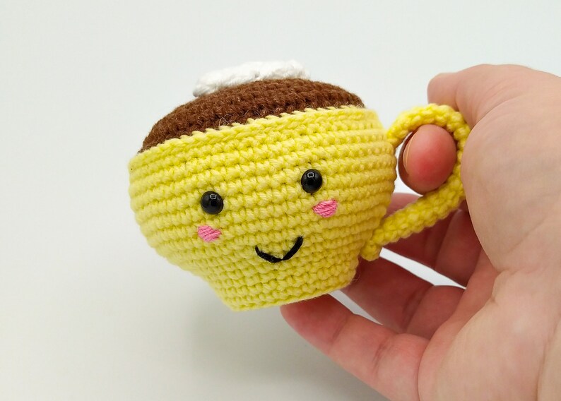 May include: A crocheted yellow coffee cup with a brown top and a white cream swirl. The cup has a happy face with black eyes and pink cheeks.