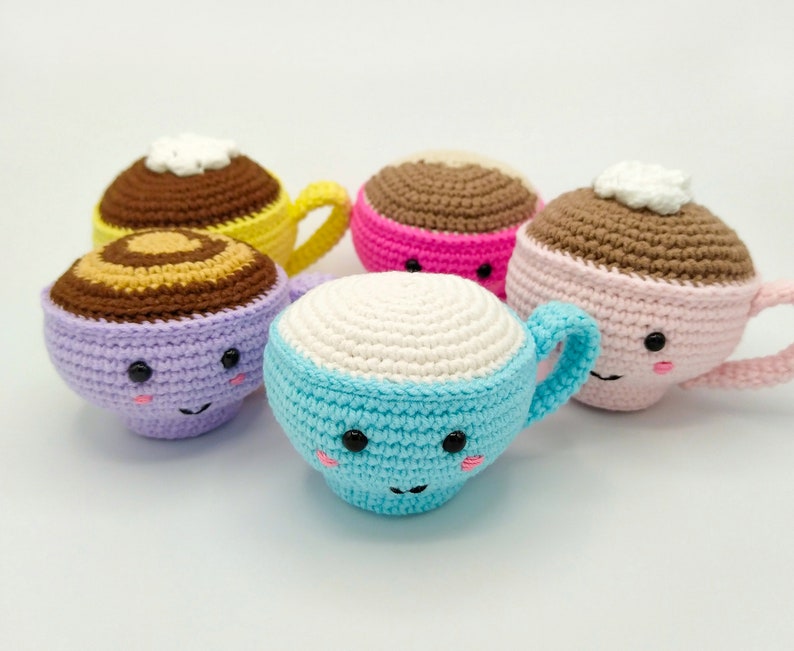 May include: Five crocheted coffee cup plushies with smiling faces. The cups are in various colors, including purple, pink, blue, and yellow. Each cup has a brown crocheted rim and a white crocheted top.