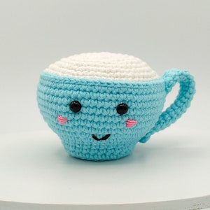 May include: A crocheted blue and white cup with a smiling face. The cup has black eyes, pink cheeks, and a black stitched smile.
