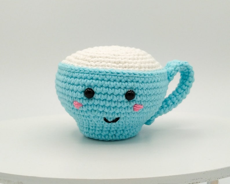 May include: A crocheted blue and white cup with a smiling face. The cup has black eyes, pink cheeks, and a black stitched smile.