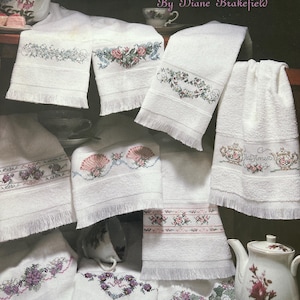 May include: A collection of white fringed towels featuring various cross-stitch floral and tea-themed patterns. The image includes the text Tea Time Towels By Diane Brakefield, along with visible ceramic teapots and teacups arranged among the textiles.