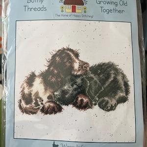 May include: Cross-stitch kit featuring a design of two dogs, with the text "Growing Old Together." The kit includes Zweigart Aida fabric, a needle, and instructions. Finished size is 10.2 inches by 10.2 inches. The brand is Bothy Threads, designed by Hannah Dale.