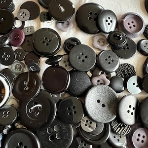 Könnte beinhalten: Eine Sammlung verschiedener schwarzer Knöpfe in unterschiedlichen Größen und Formen. Einige Knöpfe haben zwei oder vier Löcher, andere einen Schaft. Die Knöpfe bestehen aus Kunststoff und anderen Materialien, wobei einige eine matte oder glänzende Oberfläche haben.