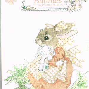 Peut inclure: Livre de patrons de point de croix intitulé "Bashful Bunnies" par Priscilla Hillman. La couverture présente une image en point de croix d'un lapin avec un petit lapin sur un objet en forme de citrouille, entouré de feuillage vert. La palette de couleurs comprend l'orange, le vert et le blanc.