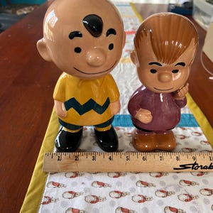 Peut inclure: Deux figurines en céramique des personnages de Peanuts, Charlie Brown et Linus van Pelt. Charlie Brown porte une chemise rayée jaune et noire et des chaussures noires. Linus porte une chemise violette et des chaussures marrons. Les figurines sont debout sur un tissu blanc avec un motif de lapins.