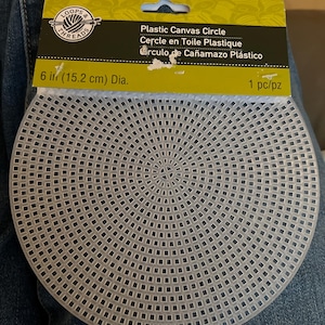 May include: A clear, circular plastic canvas sheet with a grid pattern, measuring 6 inches in diameter. The item is attached to a green and black cardboard header card labeled Loops & Threads, Plastic Canvas Circle, 6 in (15.2 cm) Dia., 1 pc/pz.