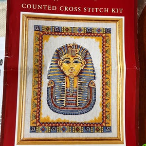 May include: Counted cross stitch kit featuring a colorful image of Tutankhamun, the Egyptian pharaoh, with the text "Tutankhamun" and "Anchor Counted Cross Stitch Kit" on the packaging. The kit includes 14 count linen blend Aida, stranded cotton, a chart, full instructions, and a needle. Size: 12" x 10".