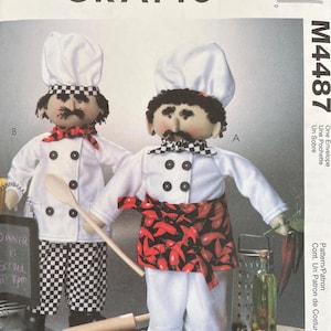 May include: McCall's Crafts M4487 sewing pattern envelope featuring two fabric chef dolls. One doll wears a white chef hat, white jacket with black buttons, and black and white chequered trousers. The other wears a white hat, jacket, and a red chilli pepper print apron.