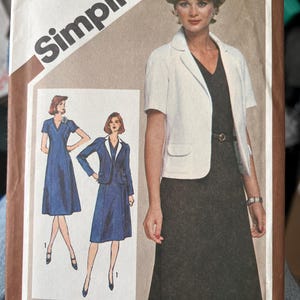 Può includere: Una busta per cartamodello Simplicity, numero 9853, taglia 18 1/2 half-size. La copertina presenta un'illustrazione di un abito scuro e una giacca bianca a maniche corte. Il testo include 'Simplicity', '9853', '$2.50', 'SIZE 18 1/2'.