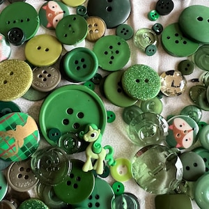 Könnte beinhalten: Eine Sammlung grüner Knöpfe in verschiedenen Formen und Größen, von Olivgrün bis Smaragdgrün. Einige Knöpfe sind mit Glitzer versehen, andere weisen Tiermotive auf. Eine kleine grüne Ponyfigur ist ebenfalls vorhanden.