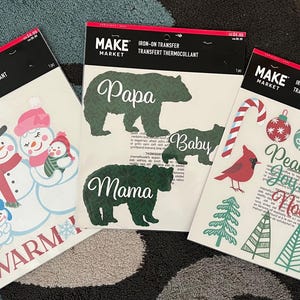 May include: Three iron-on transfer sheets from Make Market. One features a snowman family with the word "WARM". Another has bear silhouettes with the words "Papa", "Mama", and "Baby". The third has Christmas-themed graphics and the words "Peace, Joy, Noel".