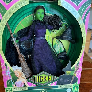 Puede incluir: Una muñeca Elphaba de Wicked con un vestido morado texturizado, capa negra, escoba, sombrero puntiagudo negro y un bolso pequeño negro. El empaque incluye el logotipo de Wicked, una imagen de Glinda y una imagen de Elphaba sosteniendo una escoba.