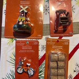 May include: Four packages of miniature decorations for a dollhouse or miniature village. The packages feature a windmill, a rocking chair, a bicycle with a flower basket, and hay bales. Each package is labeled with the brand name Ashland and the price.