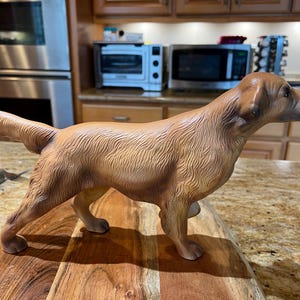 May include: A detailed, brown ceramic dog figurine in a standing pose. The dog has a smooth, glossy finish and is positioned on a wooden cutting board. The figurine is a decorative item, likely for home decor.