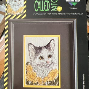 May include: A framed cross-stitch kit featuring a kitten with a white and gray coat, surrounded by yellow flowers. The kit is titled "Kitten Called Patches" and includes a 5"x7" design that fits an 8"x10" frame. The kit is designed by Sydney McIntosh.
