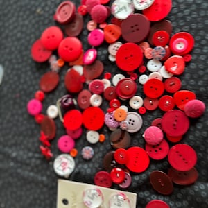 Könnte beinhalten: Eine Sammlung roter, rosa und weißer Knöpfe in verschiedenen Größen und Formen. Einige Knöpfe sind rund, andere oval, und einige haben ein Blumenmuster. Die Knöpfe liegen auf einer dunklen, gemusterten Oberfläche.