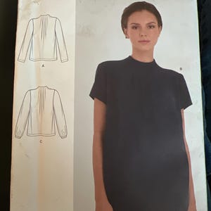 May include: A Simplicity sewing pattern envelope S9231 U5 for sizes 16-24. The white paper packaging features a dark navy short-sleeved mock neck top with front pleats, alongside line drawings of two additional long-sleeved top variations.