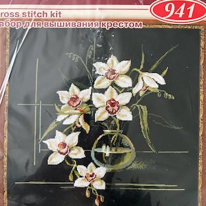May include: RIOLIS cross stitch kit 941 featuring a floral design of white orchids with dark centers in a glass vase against a black background. The packaging displays the finished size as 15 3/4 inches by 15 3/4 inches, 40 by 40 cm.