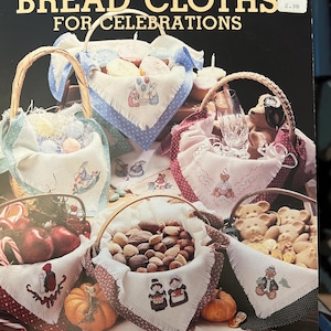 May include: A leaflet titled "Bread Cloths for Celebrations" features various baskets filled with food and decorated with embroidered cloths. The cloths have different holiday-themed designs, including bunnies, pilgrims, and a witch. The cover is black with gold lettering.