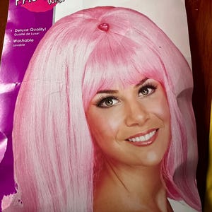 Puede incluir: Peluca rosa con estilo de volteo, empaquetada en un fondo rosa y morado con el texto "FLIP PINK WIG" y "Perruque Rose".